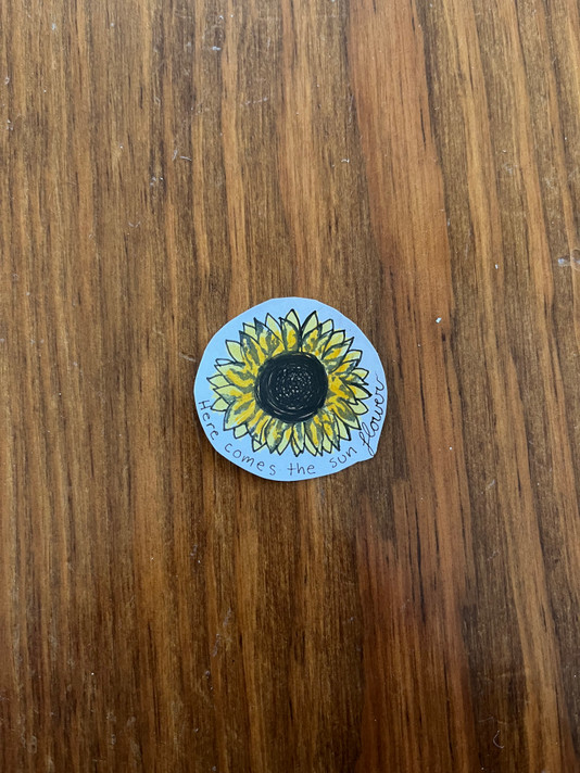A colored handmade recyclable "here comes the sunflower" sunflower sticker on a wooden floor
