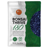 A professional-looking, resealable stand-up pouch of Bonsai Thrive 180, a time-release fertilizer specifically formulated for the specialized needs of bonsai trees. 

The packaging design is split vertically. The left side has a clean, textured white background featuring the product name "BONSAI THRIVE 180" in bold, black lettering beneath an orange circular logo of a stylized crane. Below the product name, several key horticultural benefits are listed: "Long-Lasting Nutrient Supply," "Low Risk of Over-Fertilization," "NPK 19-5-9 in Every Granule," and "Improved Root Development." 

The right side of the bag is a deep forest green adorned with delicate, light-green line-art illustrations of leaves and branches. A large, clear, crescent-shaped window on this side reveals the actual fertilizer inside: small, uniform, spherical granules in shades of deep indigo and light blue. 

At the bottom of the pouch are icons for "100% Satisfaction Guaranteed" and a green checkmark indicating the product is "Ready to Use." The weight is listed as Net Wt. 10 oz (280 g). The overall image conveys a high-quality, specialized maintenance product for bonsai enthusiasts.