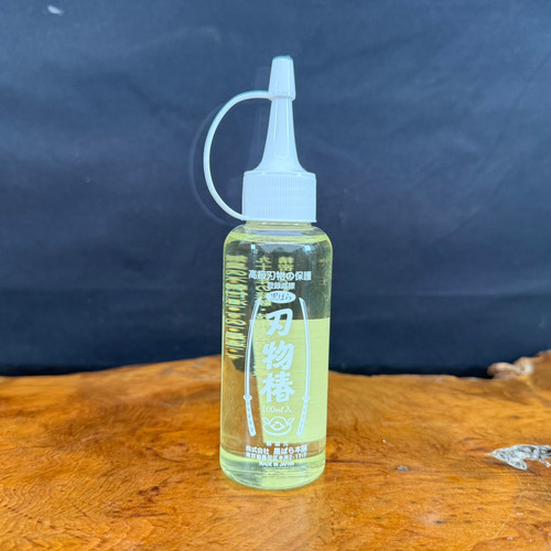 A 100ml clear plastic bottle of Kurobara Camellia Oil, also known as Hamono Tsubaki, stands vertically on a polished natural wood surface with a prominent grain. The bottle contains a light yellow oil and is topped with a white screw-on cap featuring a long, narrow applicator tip for precise tool oiling. The label features Japanese kanji, including the large characters "刃物椿" (Hamono Tsubaki), framed by illustrations of traditional Japanese swords. The background is a solid, dark-colored fabric, which makes the bottle stand out. This specialized oil is used for the maintenance and rust protection of high-quality bonsai tools and carbon steel blades.