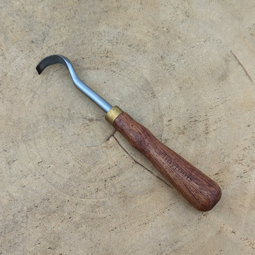 A high-quality Tinyroots bonsai carving tool is displayed diagonally on a rustic wooden slab. This specialized hand tool, used for creating intricate jin and shari details in bonsai trees, features a dark, ergonomically shaped wooden handle with the "Tinyroots" brand name engraved along its side. A gleaming brass ferrule secures the handle to a strong, curved metal neck. At the tip, the tool has a sharp, hooked blade designed for precision carving and stripping bark. The tool rests on a light-colored wood cross-section, which reveals natural grain patterns and growth rings, highlighting the tool's intended use in fine woodworking and horticultural art.