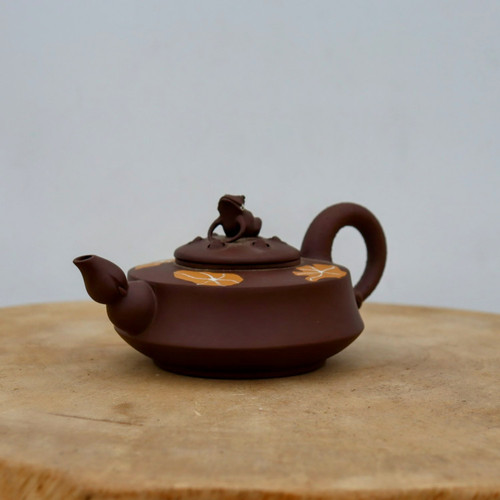 A detailed photograph of a handmade Yixing teapot, showcasing traditional Chinese purple clay (zisha) craftsmanship. This squat, dark brown teapot features a sharp angular body and is decorated with light orange floral patterns on its shoulder. The lid is uniquely accented with a small, meticulously sculpted frog figurine acting as a knob. With a short spout and a sturdy C-shaped handle, the teapot sits elegantly on a rustic wooden base, representing the aesthetic of a classic tea ceremony accessory.