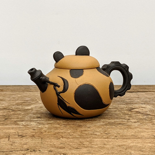A meticulously handcrafted Yixing clay teapot with a playful panda motif, resting on a weathered wooden surface against a neutral background. The teapot's rounded, sandy-tan body is adorned with bold, matte-black patches that mimic the distinctive markings of a giant panda. 

The lid is subtly shaped like a panda's head, complete with two small, rounded black ears that serve as functional grips. Adding to the nature-inspired theme, the spout is modeled after a black bamboo stalk, featuring a small, etched bamboo leaf detail that extends elegantly onto the teapot's side. The handle is a striking circular shape with a decorative, notched or scalloped outer edge, also finished in a contrasting matte black. This artisanal piece showcases the traditional craftsmanship of Yixing pottery, blending functional design with charming, animal-themed artistry suitable for a tea ceremony or as a unique home decor accent.