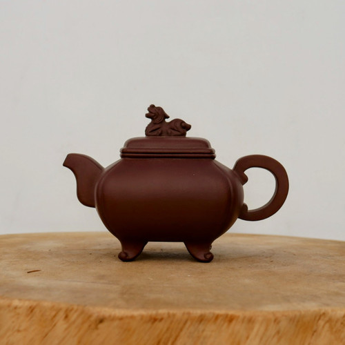 An image of a traditional Yixing clay teapot, a classic example of Chinese functional art, is shown from a side profile. The teapot is crafted from a deep, matte reddish-brown Zisha clay, prized for its ability to enhance the flavor of tea over time. 

The body of the teapot features a soft, rectangular form with rounded edges, elegantly supported by four delicate feet that curl slightly outwards at the base. A matching lid sits perfectly on the rim, topped with a finely sculpted decorative knob in the shape of a reclining mythical creature, such as a shishi or lion dog. A short, upward-curving spout is balanced on the opposite side by a smooth, rounded handle, designed for both aesthetic harmony and ease of use. 

Set upon a thick, natural wooden surface against a clean, off-white background, the teapot’s smooth yet earthy texture is highlighted by soft, even lighting. This piece exemplifies the craftsmanship of Yixing pottery, making it a sought-after item for tea enthusiasts and a beautiful accessory for bonsai displays or traditional tea ceremonies.