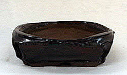A small, handmade dark-glazed ceramic pot with an irregular, organic shape, measuring approximately 4.25 inches wide and 1.25 inches deep. This rustic, shallow container features a lumpy, uneven rim and a deep espresso-brown finish with a subtle sheen, designed specifically for kusamono or accessory plantings in bonsai displays. The pot sits on a neutral, off-white background, highlighting its hand-formed texture and low-profile silhouette.