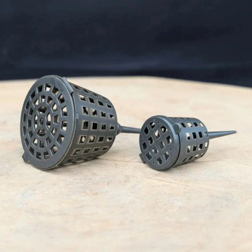 Two dark gray plastic bonsai fertilizer cups, one medium and one small, rest on a light tan, textured stone surface against a dark, blurred background. 

These reusable baskets are designed with a mesh-like cage body featuring small square openings to hold organic fertilizer pellets while allowing water to flow through for a steady, slow-release nutrient delivery. Each cup features a hinged, circular lid with a radial spoke design that snaps shut to protect the pellets from being scattered by birds or pests. At the base of each basket is a long, tapered plastic spike, which is used to securely anchor the device into the bonsai’s soil mix. 

The larger "medium" cup on the left is shown tilted forward to reveal the intricate lid design, while the "small" cup on the right is shown in profile, highlighting its compact size and sharp spike. The durable, UV-stable plastic is built for year-round outdoor use, keeping the soil surface tidy for professional bonsai displays.