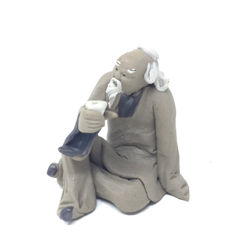 A detailed studio photograph of a traditional Chinese mud man clay figurine, commonly used as a decorative bonsai tree accessory. The small sculpture depicts an elderly sage with a wise expression, featuring a long, textured white beard and a matching handlebar mustache. He has wisps of white hair on the sides of his head and a bald top. 

The man is sitting in a relaxed, cross-legged pose. He is dressed in a simple, loose-fitting grey robe with dark blue accents visible at the sleeves. In his right hand, he holds a small, white tea cup up towards his face as if he is about to take a sip, while his left arm is extended behind him, resting on the ground for support. The figurine has a handcrafted, rustic aesthetic with visible textures in the clay, set against a solid white background.
