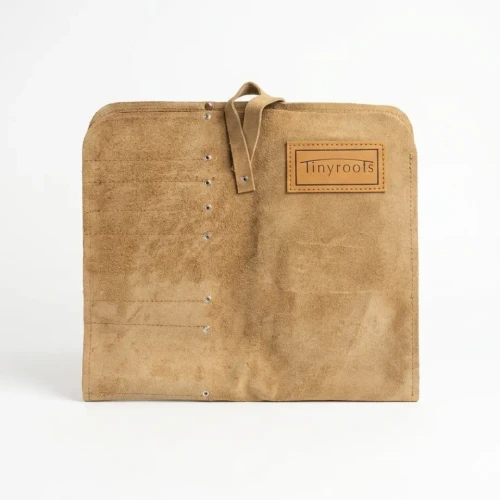 A front-view studio photograph shows a Tinyroots suede tool roll bag, likely for bonsai or artisan tools, laid flat against a solid white background. 

The bag is crafted from a light tan or camel-colored suede with a soft, brushed texture. It has a rectangular shape with slightly rounded top corners. On the left side, several vertical columns of stitching create narrow pockets for individual tools. These pockets are reinforced with small, silver metal rivets at the top and bottom of each seam. 

In the top center of the bag, a thin suede strap is attached, intended to wrap around and secure the bag once it is rolled up. On the upper right side, a rectangular, smooth leather patch is neatly stitched to the surface. It features the brand name "Tinyroots" embossed in a clean, serif font inside a thin rectangular frame. The entire piece is finished with matching perimeter stitching, giving it a durable and professional appearance.