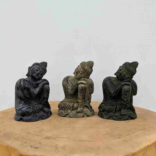 A row of three small, handmade "Relaxing Buddha" statues sits on a thick, light-brown wooden slab against a plain white background. Each figurine is intricately detailed, showing a serene Buddha in a resting pose with his head tilted and resting on crossed arms atop a single raised knee. 

The statues display a range of weathered, stone-like finishes: the one on the left is a dark charcoal grey, the center one is a lighter sandy beige, and the one on the right is a deep mossy green with a subtle patina. Their detailed features, including the traditional coiled ushnisha hair and flowing robes, highlight their craftsmanship. These peaceful figurines serve as calming decorative accents, perfect for adding a touch of Zen to a bonsai garden display or home interior.