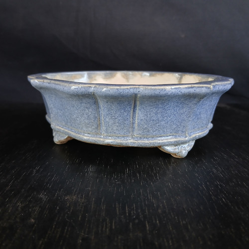 A side-profile view of a 7-inch glazed Yixing bonsai pot showcasing a mottled periwinkle blue finish. The pot's rectangular form features softened, rounded corners and sides that flare slightly outward from the base. Decorative vertical panels or flutes adorn the exterior, framed by subtle horizontal ridges near the top rim and the base. The vessel sits on four small, molded decorative feet. The interior of the pot reveals a pale, unglazed ceramic surface. The entire piece is photographed against a dark, minimalist background on a black wood-grained surface, highlighting the delicate speckled texture and cool tones of the blue glaze.