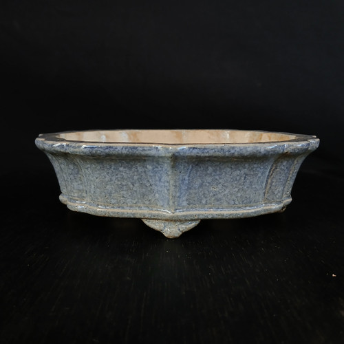 A detailed side view of a shallow, glazed Yixing bonsai pot (model YX1030-1B) set against a solid black background. The pot features a mottled, speckled glaze in shades of dusty blue and grey, with warm tan and cream tones peeking through, especially around the inner rim, giving it a textured, stone-like appearance. Its body is shaped into a lobed oval with elegant vertical ridges that create a series of subtle panels around its exterior. The pot has a slightly flared rim and is supported by several small, decorative "cloud" style feet integrated into the base. It sits on a dark, textured wooden surface, which provides a clean and professional presentation of this 7-inch ceramic container designed for bonsai trees.