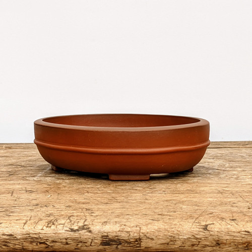 A high-angle, eye-level shot of a shallow, oval unglazed bonsai pot crafted from reddish-brown terracotta clay. The pot features a prominent horizontal decorative band or ridge that encircles the midpoint of its exterior wall, adding a subtle touch of traditional craftsmanship. The top rim is slightly thickened and rounded, while the base is supported by four small, blocky rectangular feet that elevate the pot slightly. The clay has a smooth, matte texture with a uniform warm, earthy tone. The pot is positioned in the center of a rustic, weathered wooden tabletop with visible grain and aged details, set against a clean, minimalist off-white background. This type of shallow oval pot is typically used for wide-spreading bonsai trees, such as forest plantings or deciduous species, where a low-profile container enhances the sense of a natural landscape.