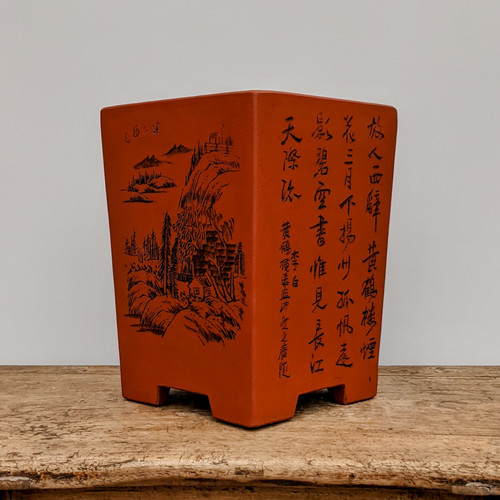 A tall, square, unglazed reddish-brown terracotta bonsai pot rests on a rustic, weathered wooden table against a plain, light-colored wall. The pot features a tapered design with four distinct block-like feet at the base. One side of the pot is adorned with a detailed black-ink engraving of a traditional Chinese landscape, illustrating rugged mountain peaks, dense pine forests, and a small pavilion nestled on a hillside. The adjacent side to the right displays several columns of elegant Chinese calligraphy etched in black ink. The earthy texture of the unglazed clay and the fine artistic details highlight its traditional craftsmanship, making it a sophisticated vessel for a bonsai display.