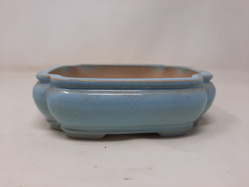 An eye-level, side-view photograph of a 7-inch glazed Yixing bonsai pot in a soft, light blue color. This shallow ceramic planter features a traditional mokkoh or scalloped rectangular shape with four rounded, lobed corners. The glossy cerulean glaze has a smooth finish with subtle dark speckles and a slightly weathered texture. 

The pot's design includes a wide, flat rim and a curved body that tapers slightly toward the base, resting on small, integrated feet. The unglazed interior reveals a natural buff-colored clay, providing a porous surface suitable for bonsai root health and drainage. The pot is presented against a plain, neutral white background, highlighting its elegant shape and serene blue hue.