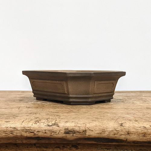 A studio photograph captures a side view of a traditional unglazed bonsai pot resting on a weathered wooden surface. The pot, crafted from dark brown ceramic with an earthy, matte finish, features a distinctive octagonal shape. Its design is characterized by a wide, flared rim at the top and a series of recessed rectangular panels on each of its eight sides, adding a subtle architectural detail. 

The pot is slightly elevated by small, block-like feet integrated into the base, which are essential for drainage. It sits on a light-toned, rustic wooden tabletop that shows natural grain and wear, providing a textured contrast to the smooth clay. The entire composition is set against a stark, solid white background, which highlights the pot's clean lines and rich, natural color, making it the central focus of the image.