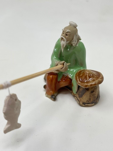 A detailed studio shot of a traditional Chinese mudman figurine, frequently used as a decorative accessory for bonsai trees. The miniature figure depicts an elderly man with a long white beard and mustache, wearing a topknot and a bright green glazed robe over orange-brown pants. He sits in a relaxed pose on a craggy, brown glazed rock, holding a simple wooden fishing rod that extends toward the viewer. A small, unglazed white ceramic fish dangles from a thin white string at the tip of the rod. To his left, a large, glossy brown conical hat rests on the rock beside him. The figurine stands against a neutral, off-white background, highlighting the contrast between the polished glaze of the clothing and the matte, naturalistic details of the face and hands. This handcrafted piece represents a serene fishing scene, intended to add a sense of scale and traditional Chinese culture to a miniature garden or indoor bonsai display.