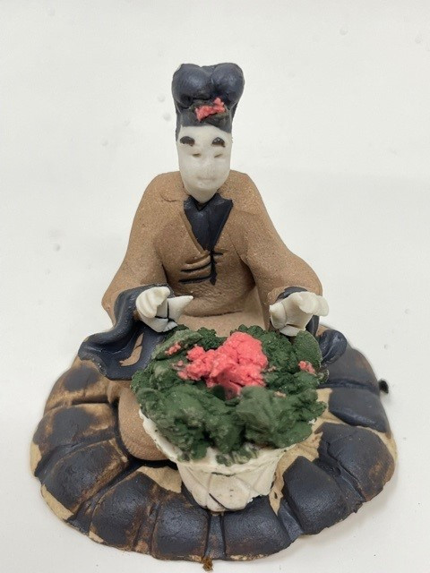 A detailed, handcrafted Chinese mudman figurine depicting a woman sitting behind a flowering pot. The miniature figure features a pale, serene face with simple painted features and dark hair styled in a traditional topknot, accented by a small red ornament. She is dressed in a tan or light brown robe with wide, dark-trimmed sleeves that have a subtle texture. Her hands are positioned as if she is carefully gesturing toward or tending to the plant in front of her.

The plant is held in a small, white, cross-hatched textured pot and consists of thick green foliage topped with vibrant pink or coral-colored flowers. The entire figurine is mounted on a circular, dark-colored base with a rough, stone-like texture and brown highlights. This rustic clay ornament, with its hand-molded characteristics, is a classic example of a Chinese mudman, often used as a decorative accent for bonsai tree displays or miniature indoor gardens to add a sense of life and tradition.