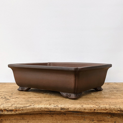 A rectangular unglazed bonsai pot crafted from dark, earthy brown ceramic is shown from a side-angle view. The pot features a prominent, slightly flared rim and four ornate decorative feet at each corner, embellished with a delicate scrollwork or "cloud" pattern. Its matte, unglazed finish gives it a natural, traditional appearance. The bonsai pot sits centered on a weathered, light-colored wooden surface, likely a rustic bench or table, against a clean, minimalist off-white background that emphasizes the pot's form and color.
