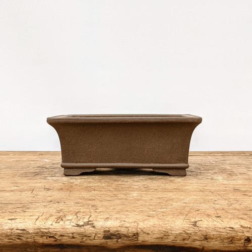 A side-profile view of a rectangular, unglazed brown ceramic bonsai pot resting on a weathered, light-colored wooden tabletop. The pot features a minimalist design with a subtly flared rim and four small, integrated corner feet that lift it slightly off the surface. Its earthy, fine-textured brown clay finish is characteristic of traditional unglazed bonsai containers, which are often favored for coniferous tree species like pines and junipers to create a balanced, natural aesthetic. The composition is centered against a clean, plain white background, highlighting the pot's elegant lines and professional craftsmanship.