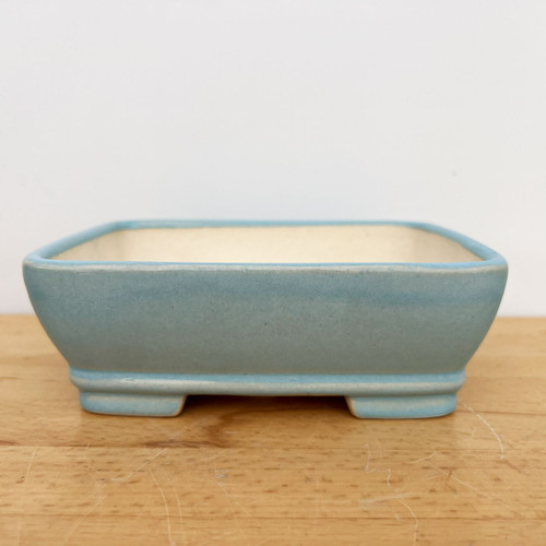 A side profile view of a 7-inch light blue glazed Yixing bonsai pot resting on a light-colored wooden table against a plain white background. This rectangular ceramic pot features a smooth, light blue glaze with delicate tonal shifts and rounded corners. It has a slight outward taper from its base to a thick, rounded rim. The pot sits on four small, rectangular feet, and its interior reveals a pale, off-white unglazed ceramic finish. This elegant and functional container is designed for small to medium-sized bonsai trees.