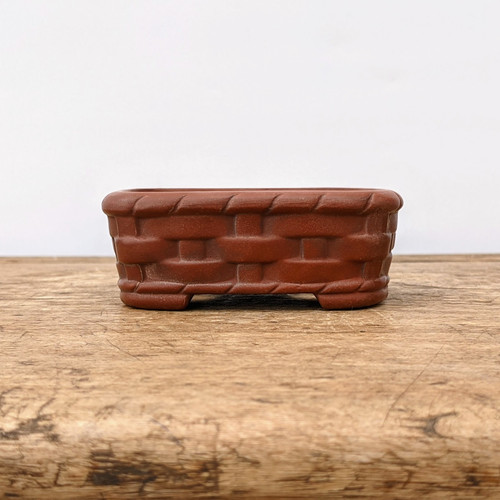 A close-up, eye-level photograph of a small, rectangular Japanese Tokoname bonsai pot crafted by the renowned Bigei kiln. The pot is made from a rich, unglazed reddish-brown clay and features an intricate, molded basket-weave texture on its exterior. Horizontal bands appear to weave in and out of vertical supports, giving the piece a rustic and traditional aesthetic. Both the top rim and the base of the pot are adorned with a decorative, twisted-rope border. The pot rests on small, integrated corner feet and is positioned on a weathered, light-colored wooden surface against a clean, off-white background. This piece highlights the exquisite craftsmanship and natural beauty of authentic Japanese bonsai pottery.