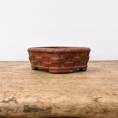 A small, rectangular Japanese Tokoname bonsai pot by the Bigei kiln sits on a rustic wooden surface. Crafted from unglazed, reddish-brown clay, the pot features a detailed, hand-sculpted basketweave pattern across its exterior. The upper rim and the base are finished with a raised, rope-like decorative border that complements the woven texture. The pot has rounded corners and sits elegantly on four small, integrated corner feet. It is photographed at eye level against a clean, minimalist white background, highlighting its rich earthy tones and artisanal craftsmanship.