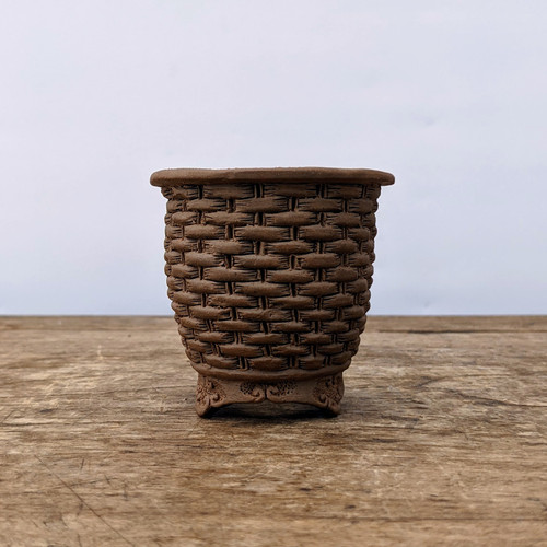 A professional studio photograph displays a small, round Japanese Tokoname bonsai pot, expertly handcrafted by the artist Bigei. The pot is made from unglazed, dark terracotta-colored clay, featuring an intricate and deeply textured basket-weave or wicker pattern that covers the entire exterior body. 

The pot has a smooth, slightly flared rim at the top and tapers gently toward the base. It stands on four small, decorative feet that are carved with delicate scroll or cloud-like motifs, providing a touch of elegance to its rustic aesthetic. 

The piece is centered on a weathered, horizontal wooden surface with a prominent grain and minor scuffs, suggesting an organic, natural setting. The background is a solid, neutral white, which makes the warm brown tones and fine details of the ceramic work stand out clearly. The even lighting highlights the three-dimensional quality of the woven texture, showcasing the high-quality craftsmanship characteristic of traditional Japanese Tokoname pottery.