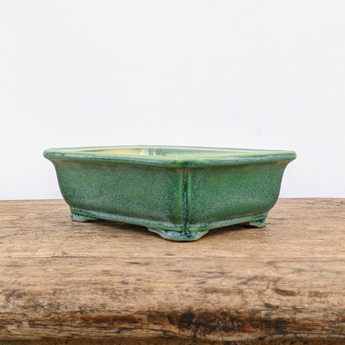 A side-angle photograph of a rectangular green glazed bonsai pot, resting on a rustic, weathered wooden table against a plain white background. The ceramic bonsai pot features soft, rounded corners, a subtle outward-flared rim, and sits on four small integral feet for drainage. The deep forest green glaze has a beautiful mottled, speckled texture, with highlights of a lighter, moss-colored green concentrated along the top edge and vertical corner lines, giving it an artisanal, hand-crafted appearance. The weathered wood surface below has prominent grain and a warm brown tone, providing a natural contrast to the smooth, glossy finish of the glazed pot.