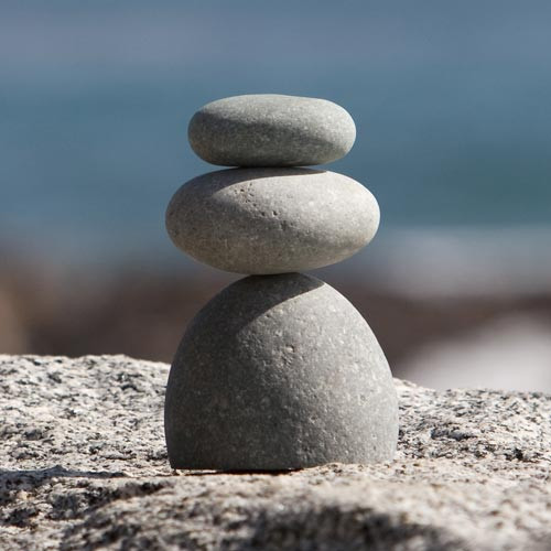 A serene close-up photograph of a triple rock cairn, consisting of three smooth, grey river stones stacked in a state of perfect balance. The stack sits upon a textured, light-colored granite surface. The bottom stone is the largest and most substantial, possessing a rounded, dome-like shape. Perched horizontally on top is a medium-sized, egg-shaped stone, which supports the smallest stone—a flattened, oval pebble—at the very top of the arrangement. 

Bright, natural sunlight coming from the upper right illuminates the scene, highlighting the fine, speckled mineral texture of the stones and casting soft, gentle shadows on their left sides. The background is a beautifully blurred coastal landscape, featuring a calm blue sea meeting a soft horizon under a clear sky, which enhances the image's overall sense of peace and tranquility. This arrangement of stones, often used in zen gardens and as accents for bonsai, symbolizes mindfulness, harmony, and spiritual balance.