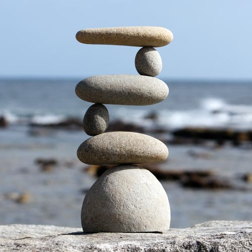 A serene medium-sized rock cairn stands perfectly balanced against the backdrop of a blurred coastal scene. The stack consists of six smooth, light-colored river stones arranged in an intricate "side2side" or zig-zag pattern. It begins with a wide, dome-shaped base stone that supports a flat, oval rock. Above this, the weight shifts laterally using smaller rounded stones as pivot points for alternating long, flat rocks that extend to the left and right. 

The stones' natural textures of grey and tan are highlighted by bright daylight. In the background, the soft focus reveals a deep blue ocean with white foamy waves and a clear sky, emphasizing the cairn's peaceful and meditative presence. This artistic arrangement of stacked rocks serves as a symbol of stability and zen-like balance, often used as a decorative accent in outdoor gardens or alongside bonsai trees.