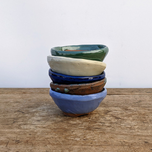 A vertical stack of five small, hand-crafted glazed ceramic pots sits in the center of a rustic, weathered wooden tabletop. The pots, ideal for miniature bonsai trees or succulents, feature unique, slightly irregular shapes and varied finishes. 

From top to bottom, the colors include:
*   A glossy, forest green pot with a wavy rim.
*   A matte, off-white or cream-colored pot.
*   A deep, shiny cobalt blue pot.
*   An earthy, textured terracotta brown pot with subtle green glaze drips.
*   A soft, matte periwinkle blue pot at the base.

The background is a clean, solid off-white wall, which emphasizes the artisanal textures and vibrant colors of the ceramic stoneware. Each pot has a shallow design typical of succulent or bonsai containers.