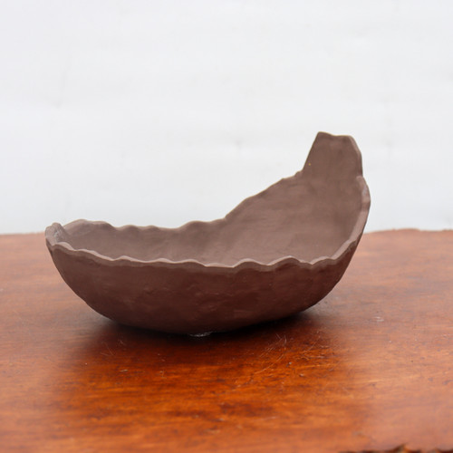 A side view of a small, unglazed Japanese ceramic planter in a rich, earthy chocolate-brown color. This specialty bonsai pot features an organic, asymmetrical shape similar to a hollowed-out seed pod or an open shell, with a tall, angled peak on one end. The top rim is meticulously finished with a wavy, scalloped pattern, and the exterior has a slightly textured, hand-molded quality. The pot rests on a warm, polished wooden tabletop with a visible grain, set against a plain, soft-white background that emphasizes its unique form and earthy texture.