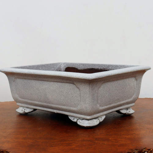 A high-quality Yixing glazed bonsai pot in a rectangular shape with rounded corners. The pot's exterior is covered in a beautiful light gray or off-white crackle glaze, creating an intricate network of fine lines that give it a weathered, antique character. The design features recessed panels on the sides and a prominent flared rim. It is elevated by four ornate, scroll-like feet that add a touch of traditional elegance. The pot is photographed from a slight side angle, resting on a polished, dark reddish-brown wooden surface against a clean, neutral white background, highlighting its craftsmanship and subtle coloration. This ceramic pot is an example of refined Yixing pottery, designed to complement a wide variety of bonsai tree styles.