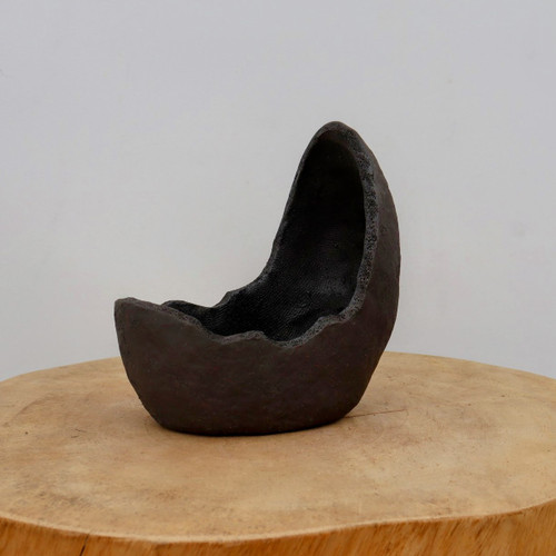 A dark charcoal, unglazed ceramic succulent planter with a rough, earthy texture sits centered on a round, light-colored wooden pedestal against a plain white wall. The pot has a unique, organic shape reminiscent of a broken geode or a hollowed-out stone. One side curves upward into a tall, pointed peak, while the opposite side is lower with a jagged, irregular rim that mimics a natural break. 

The deep interior of the planter is dark and spacious, designed to accommodate soil for a small bonsai tree or succulents. The matte, deep-brown-to-black finish and primitive, asymmetrical design of this unglazed bonsai pot create a strong naturalistic aesthetic, making it an ideal decorative piece for minimalist, wabi-sabi, or rustic-style home decor. The lighting highlights the pitted and uneven surface of the clay, emphasizing its handcrafted quality.