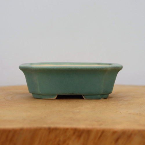 A side-view photograph of a rectangular glazed ceramic bonsai pot in a soft, matte seafoam green or teal color. The pot features slightly flared upper edges and distinctive indented corners, giving it a refined, octagonal profile. Its glaze shows subtle, earthy undertones along the rim and base, adding a touch of antique character to the piece. It rests on a thick, light-colored natural wood surface against a minimalist off-white background. This durable and stylish ceramic pot is designed specifically for bonsai trees, offering a perfect blend of form and function for both indoor and outdoor displays.