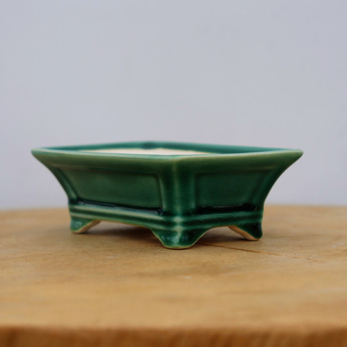 A side-view photograph shows a small, rectangular glazed ceramic bonsai pot in a vibrant deep forest green. The pot features a glossy finish with subtle crazing textures throughout the glaze. Its design includes flared sides, a distinct rim, and recessed rectangular panels on the front and sides. The container stands on four small, elegant feet that elevate the base. It is resting on a light-toned, natural wood surface against a minimalist, neutral light gray background that makes the rich green color of the ceramic pop. This miniature bonsai pot is a classic choice for small trees, offering a traditional aesthetic for indoor or outdoor displays.