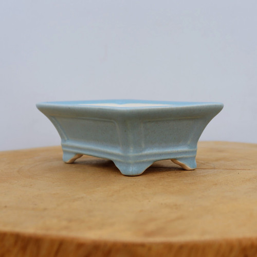 A side-view photo of a small, rectangular light blue glazed bonsai pot resting on a circular natural wood cross-section. The pot has an elegant, flared profile, widening from the base to the top rim. It features four decorative cloud-style feet and a subtle inset rectangular panel on its sides. The glaze is a soft, uniform robin's egg blue with a smooth finish. The wooden surface provides a warm, organic contrast to the cool ceramic tones, set against a simple, pale gray background, highlighting the pot's craftsmanship as a piece of functional bonsai art.