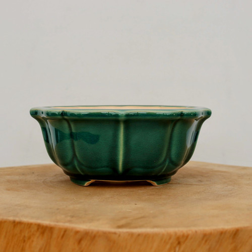 A close-up, eye-level photograph of a round, glazed ceramic bonsai pot resting on a circular cross-section of a light-colored wooden log. The pot is finished in a rich, deep emerald green glaze with a delicate crackle texture throughout. Its sides are elegantly lobed or scalloped, creating a series of vertical ridges and curves that give the vessel a floral or melon-like appearance. A faint, dark-green drip or brushstroke is visible on the left side of the pot's body. The pot has a slightly flared rim that follows the undulating pattern of the lobes. It sits on small, built-in feet, and the unglazed, pale-beige interior is partially visible at the top edge. The background is a clean, neutral off-white wall, emphasizing the pot's vibrant color and artisanal design.