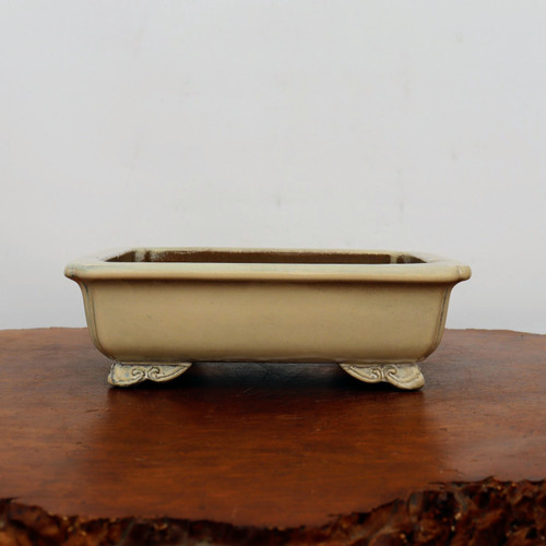 An eye-level, side-profile photograph of an empty, rectangular ceramic bonsai pot with a soft cream-colored glazed finish. The pot is positioned in the center of the frame, resting on a polished, warm-toned wood surface against a minimalist white background. 

The container features a smooth, matte cream glaze with subtle, dark weathered accents along the corners and the underside of the flared top rim, giving it a vintage or antiqued aesthetic. It is elevated by four decorative scroll-style feet, often referred to as "cloud feet," which feature ornate carved details. The shallow design and neutral color palette make this pot a versatile vessel for traditional bonsai tree displays.
