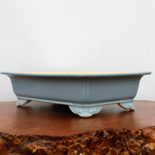A side-profile view of a rectangular, light blue glazed ceramic bonsai pot resting on a polished, dark reddish-brown wooden stand. The pot features a smooth, uniform powder-blue glaze with a wide, slightly flared rim at the top. It is supported by four decorative, scroll-carved "cloud feet" that lift the base of the pot off the surface. The thin edges of the rim and the decorative feet reveal a hint of the pale, cream-colored ceramic body beneath the glaze. The wooden base has a glossy finish on top and a textured, rustic "live edge" in the foreground. The entire scene is set against a clean, minimalist off-white background, highlighting the elegant design and craftsmanship of this ornamental bonsai tree container.