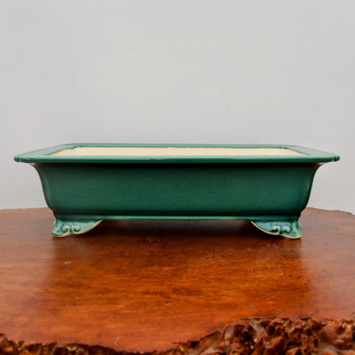 A side-view, eye-level photograph showcases a rectangular glazed bonsai pot in a deep, lustrous emerald green. The ceramic surface is finished with a fine, intricate crackle glaze that adds texture and depth to the vibrant color. The pot's shallow rectangular form features a wide, slightly flared top rim, which reveals a clean, off-white interior. At each bottom corner, the pot is elevated by elegant, scrolled decorative feet, adding a traditional touch to the design. It rests upon a polished, warm-brown wooden bonsai display stand with a visible grain and a rustic, textured live edge at the bottom right of the frame. The entire scene is set against a neutral, soft-gray background, creating a serene and professional presentation of high-quality bonsai pottery.