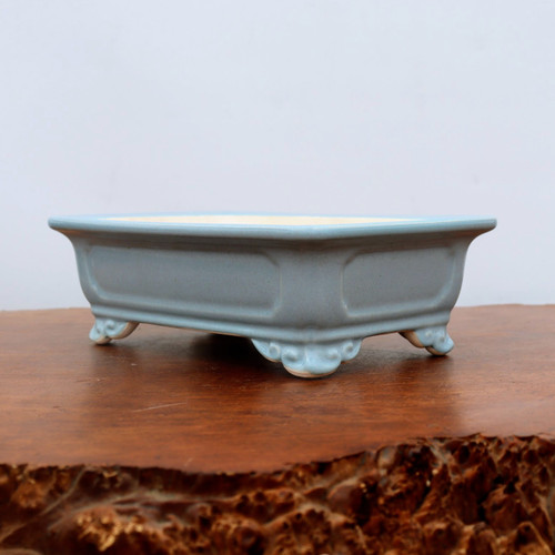 A side profile of a light blue glazed ceramic bonsai pot resting on a rustic, dark wood stand. The rectangular pot features slightly flared edges at the top and indented side panels that create a subtle decorative pattern. It is elevated by four ornate, cloud-style (ruyi) feet. The glaze is a soft, pale blue with a smooth finish, and the unglazed light-colored interior is visible at the very top rim. The background is a clean, off-white wall, emphasizing the pot's elegant silhouette and traditional design.