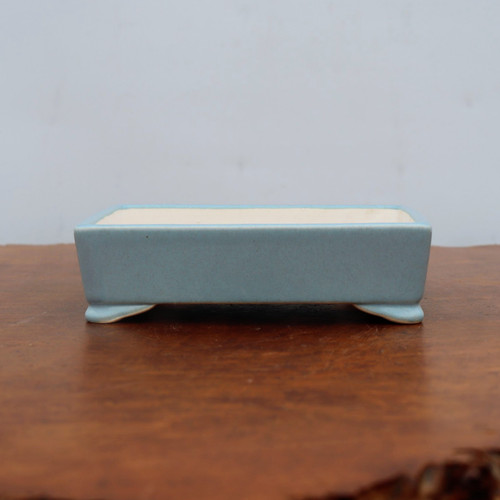 A rectangular, glazed ceramic bonsai pot in a soft, light blue shade sits on a dark, polished wooden surface. The pot features a matte, slightly mottled blue finish on its exterior, while the interior is a contrasting off-white or cream color. The rectangular form is defined by clean, straight lines and sharp corners, supported by four small, elegantly flared feet at each corner. The pot is centered in the frame, with the rich texture of the wooden table in the foreground and a plain, neutral-toned wall in the background, highlighting the container's simple and classic design.
