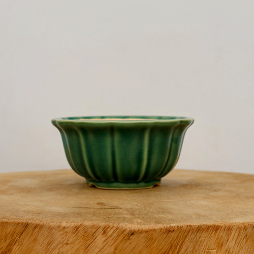A small, glazed green ceramic bonsai pot with a fluted, scalloped design is centered on a thick, circular wooden slab against a neutral off-white background. The pot features a vibrant emerald green glaze with subtle tonal variations that highlight its vertical ridges and elegant, rounded shape. It has a slightly flared rim and sits on a low, integrated base, providing a sturdy container for small bonsai trees or succulents. The smooth, shiny finish of the glazed ceramic contrasts with the natural, rustic texture of the wooden surface below, creating a minimalist and clean aesthetic.