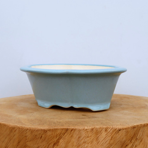 A side-view studio photograph shows a small, light blue glazed ceramic bonsai pot resting on a thick, natural wood block. This shallow, oval-shaped container features a classic scalloped "mokkou" or four-lobed design with a wide, flared rim. The exterior is finished in a smooth, uniform pale blue glaze that reflects light softly, while the interior shows the natural, off-white tone of the ceramic clay. The pot is elevated by four integrated, decorative feet with a gentle wave pattern. Set against a plain, neutral white background, the image highlights the elegant form and calming color of this bonsai container, making it an ideal choice for various bonsai tree styles.