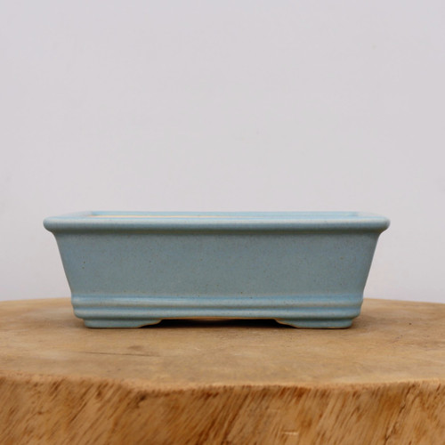 A side view of a rectangular, light blue glazed ceramic bonsai pot resting on a thick, natural wooden slab. The pot features a smooth, matte powder-blue finish with subtle flared sides and a defined upper rim. Near the bottom, decorative horizontal bands encircle the base, which is elevated by four small, integrated feet. The pot is centered on a light-colored wood slice with prominent natural grain, set against a plain, soft-white background. This elegant container is designed for displaying a small bonsai tree, offering a clean and modern aesthetic for any plant collection.