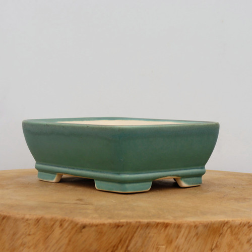 A rectangular teal green glazed ceramic bonsai pot sits atop a rustic, light-brown wooden slab against a clean, neutral white background. The pot features a soft, matte teal finish with subtle color variations, gently rounded corners, and a slight lip around the top edge. It is designed with four small, blocky feet at the base that elevate the container, revealing the unglazed pale cream ceramic underneath. This elegant and functional bonsai container is ideal for housing various bonsai tree species, providing a classic aesthetic for enthusiasts.