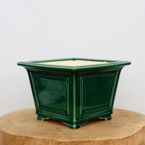 A square-shaped glazed ceramic bonsai pot in a deep, glossy emerald green. The pot features a wide, flared upper rim and sits on four integrated corner feet. Each side of the pot is decorated with a recessed rectangular panel framed by subtle raised molding, giving it a classic, structured look. The interior of the pot shows a light beige, unglazed ceramic finish. It is displayed on a thick, natural light-wood stump against a clean, off-white background.