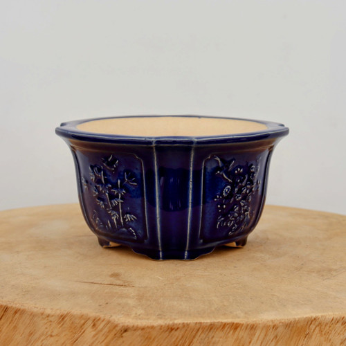 A high-gloss dark blue glazed bonsai pot sits centered on a rustic, light-colored wooden pedestal against a plain white background. The octagonal ceramic pot features a wide, flared rim and a lobed body divided into panels. Each panel is decorated with intricate embossed relief designs, including bamboo stalks and delicate floral blossoms, highlighted by the rich cobalt blue glaze. The interior of the pot shows a natural, unglazed tan ceramic finish. The piece is supported by small, integrated feet that lift the base slightly off the surface for drainage.