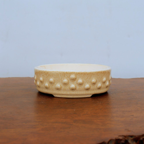 A shallow, round ceramic bonsai pot featuring a speckled cream and tan glaze with a textured hobnail design. The pot’s exterior is decorated with two staggered rows of small, raised circular studs that provide a tactile, handcrafted feel. This low-profile bonsai container, identified as model cream 2033, has a smooth interior and sits on a slightly recessed base. It is pictured resting on a polished dark brown wooden surface against a clean, neutral backdrop, highlighting its earthy tones and unique decorative finish suitable for various bonsai tree styles.