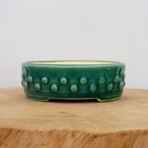A shallow, round bonsai pot featuring a vibrant emerald green glaze with a delicate crackle or crazing effect throughout. The exterior wall is uniquely decorated with two staggered rows of raised, rounded studs, resembling decorative rivets. The pot has a smooth, rounded rim and sits on a recessed footed base with small arch-shaped openings for drainage. The interior of the ceramic pot is a contrasting light cream color. It is shown resting on a thick, natural wood block with a soft-focus white background, highlighting the glossy finish and textural details of the glaze.